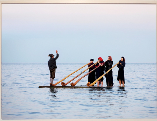 Vier menschliche Figuren (Elena Ehret, Christin Löhner, Michelle Löhner, Elias Hamann) stehen in schwarzen Gewändern nebeneinander auf einem halb im Wasser treibenden kleinen Floß und blasen in vier große Natur-Alphörner.