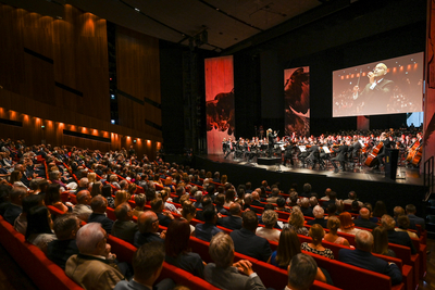 Auf diesem Bild sieht man ein Orchester, das in einem Konzertsaal spielt. Das Publikum sitzt in roten Sitzen und schaut gebannt auf die Bühne. An den Wänden sind große Projektionen zu sehen.