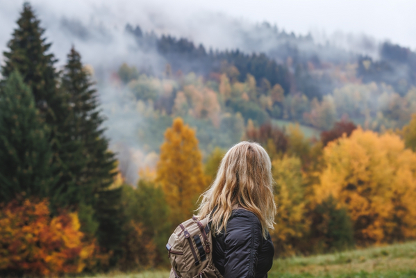 Eine junge, blonde Frau mit halblangen Haaren ist seitlich von hinten vor einer herbstlichen Hügellandschaft zu sehen. Im Hintergrund färben sich die Bäume ein, zwischen ihnen liegt Nebel in der Luft. Die Frau trägt eine dunkle Herbstjacke und einen Rucksack.