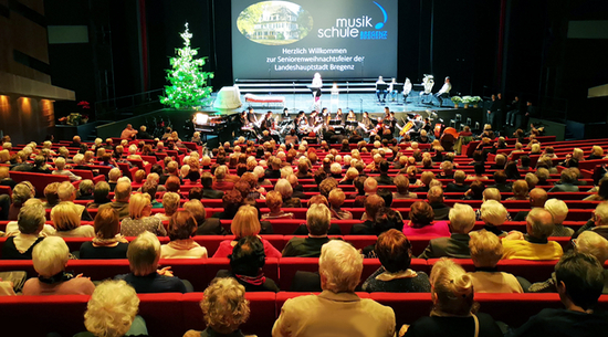 Das Bild zeigt den gefüllten Festsaal im Festspielhaus von hinten, in dem die roten Stühle von vielen Besucher:innen besetzt sind. Im Hintergrund sieht man die Bühne, auf der ein Weihnachtsbaum steht, dahinter eine große Leinwand, auf die "Musikschule Bregenz" projiziert wurde. Auf der Bühne sitzen einige Menschen auf Stühlen nebeneinander. 