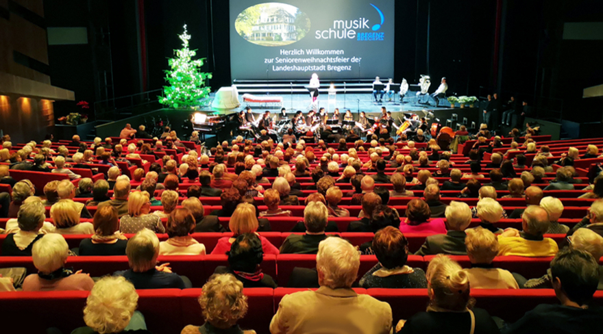 Das Bild zeigt den gefüllten Festsaal im Festspielhaus von hinten, in dem die roten Stühle von vielen Besucher:innen besetzt sind. Im Hintergrund sieht man die Bühne, auf der ein Weihnachtsbaum steht, dahinter eine große Leinwand, auf die "Musikschule Bregenz" projiziert wurde. Auf der Bühne sitzen einige Menschen auf Stühlen nebeneinander. 