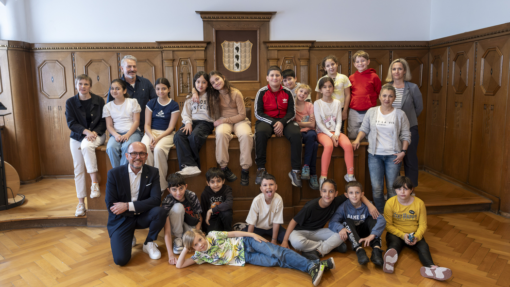 Das Bild zeigt die Schulklasse 3b der Volksschule Schendlingen beim Besuch im Bregenzer Rathaus. Die Kinder sitzen im Stadtvertretungszimmer um einen hölzernen Tisch zusammen mit ihren Lehrer:innen und Bürgermeister Michael Ritsch.