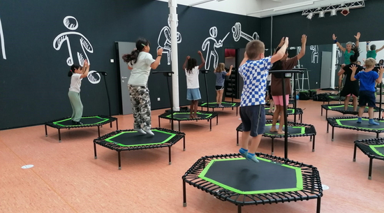 Das Bild zeigt Kinder von hinten, die in einer Turnhalle auf Trampolins springen. An der vorderen Wand sind Spiegel angebracht. Eine Lehrerin führt dort die Übungen vor.