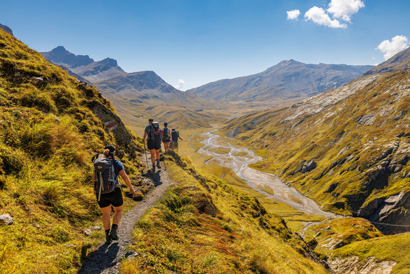 Das Bild zeigt mehrere Personen in Wanderausrüstung von hinten, die einen Weg entlang durch eine Berglandschaft wandern.