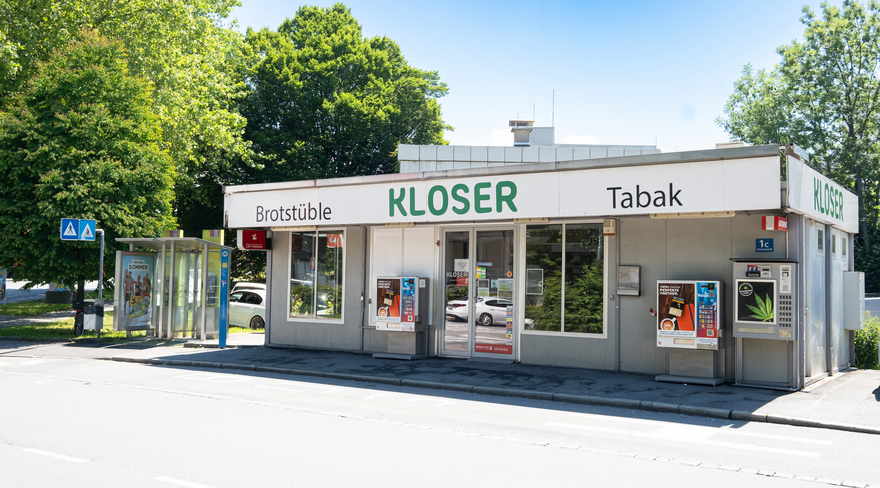 Das Bild zeigt den Kiosk in der Achsiedlung von außen fotografiert, davor der Straßenverlauf und dahinter große, grüne Bäume. 