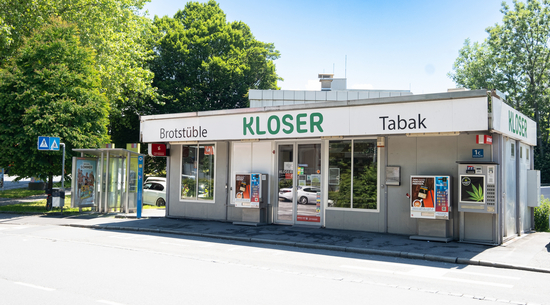 Das Bild zeigt den Kiosk in der Achsiedlung von außen fotografiert, davor der Straßenverlauf und dahinter große, grüne Bäume. 