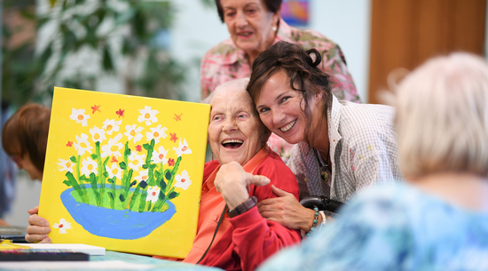 Diese Abbildung zeigt eine Dame mit einer älteren Dame zusammen, welche ein selbst gemaltes Bild hoch hält.