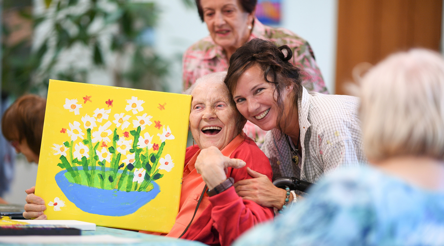 Diese Abbildung zeigt eine Dame mit einer älteren Dame zusammen, welche ein selbst gemaltes Bild hoch hält.