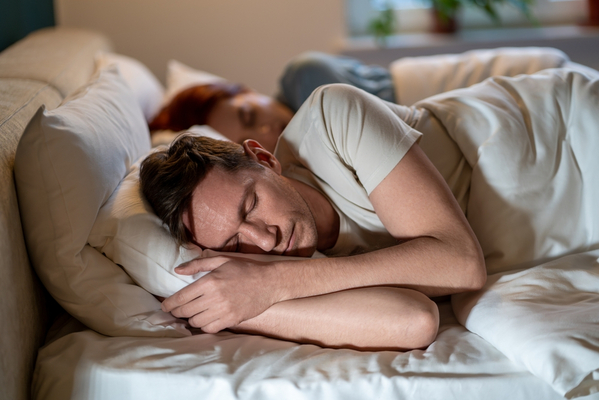 Das Bild zeigt einen Mann und eine Frau, die in einem komfortabel aussehenden, mit weißer Bettwäsche bezogenen Bett nebeneinander schlafen. Der Mann ist im Vordergrund, die Frau schläft hinter ihm.