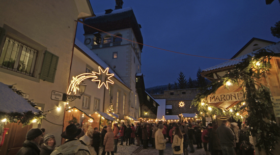 Das Bild zeigt den Weihnachtsmarkt in der Bregenzer Oberstadt in der Dämmerung. Die Weihnachtsbeleuchtung ist angeschaltet, links im Bild zum Beispiel ein beleuchteter Stern. Auf dem Marktplatz sind Marktstände angebracht, dazwischen stehen viele Menschen. Rechts im Bild steht "Maroni" auf einem Schild zu lesen.