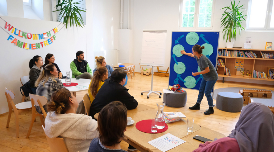 Auf der Abbildung sind mehrere Menschen auf Stühlen sitzend, sowie eine Vortagende, die im Familientreffpunkt ein Thema erörtert, zu sehen. Auf der Wand steht in bunten Buchstaben "Willkommen im Familientreff".