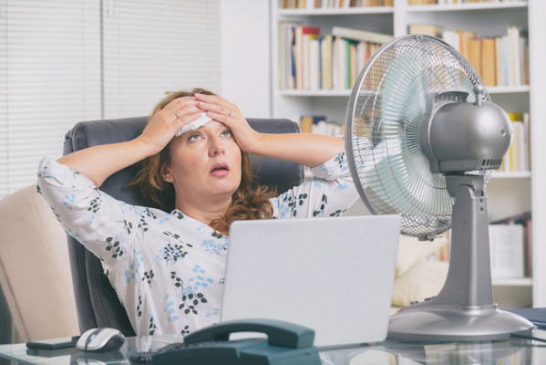 Das Bild zeigt ein Frau im Büro, die an enormer Hitze zu leiden scheint. Sie ist zurückgelehnt in ihrem Sessel und hält sich ein Taschentuch an die Stirne. Vor ihr steht ein silberner Ventilator auf ihrem Schreibtisch, daneben Laptop und Telefon. Im Hintergrund ist ein Bücherregal zu sehen.