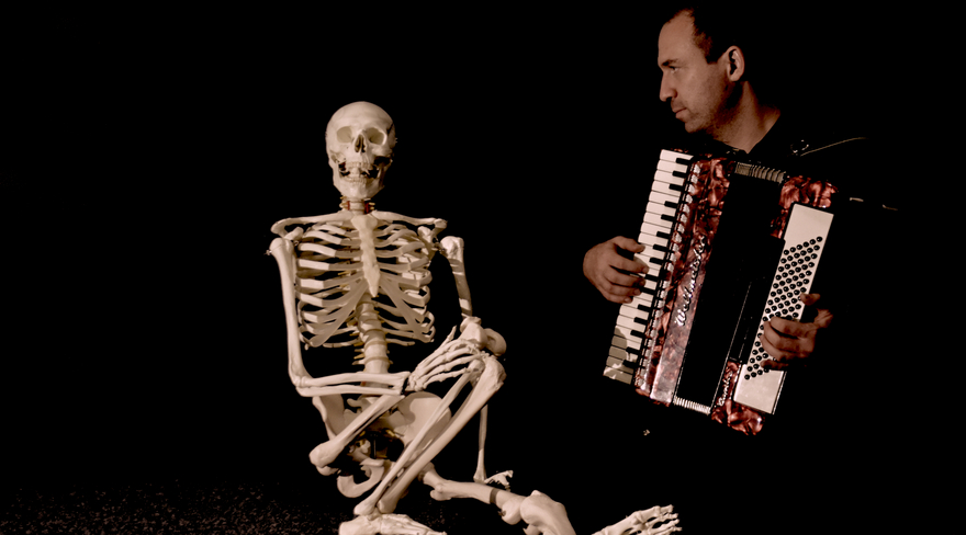Diese Abbildung zeigt einen Mann, welcher die Ziehharmonika spielt. Neben ihm sitzt ein Skelett.