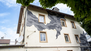 Auf dem Bild sieht man eine Hausfassade mit einem Kunstwerk aus schwarzen Linien und oben rechts sieht man die Blätter von einem Baum. 