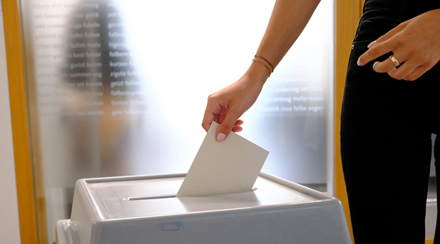 Auf dem Foto ist eine Hand mit einem Kuvert und einer Wahlurne zu sehen.