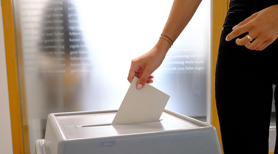 Auf dem Foto ist eine Hand mit einem Kuvert und einer Wahlurne zu sehen.