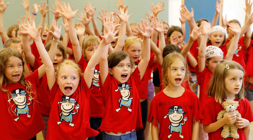 Hier wurden Kinder fotografiert, welche alle die gleichen T-Shirts tragen und ihre Hände in die Luft strecken.