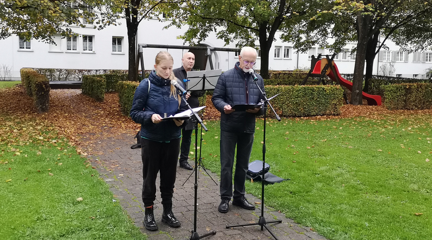 Das Bild zeigt Werner Schelling und eine Schauspielerin bei ihrer Führung und Lesung "Sie mussten sterben, weil sie anders waren". Sie stehen mit Unterlagen in der Hand vor zwei Stehmikrofonen auf einem kleinen Weg, zu einem Haus im Hintergrund führt. Neben dem Weg ist eine Wiese zu sehen.