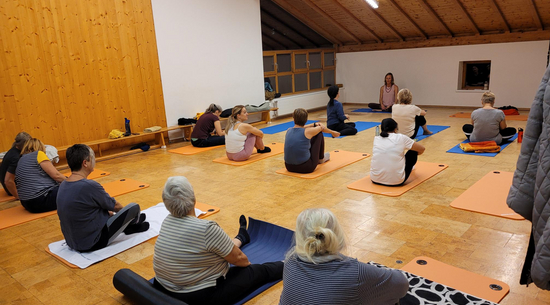 Das Bild zeigt mehrere Personen, die in einem beleuchteten Raum mit Schrägdach und Holzboden auf Yogamatten sitzen. 