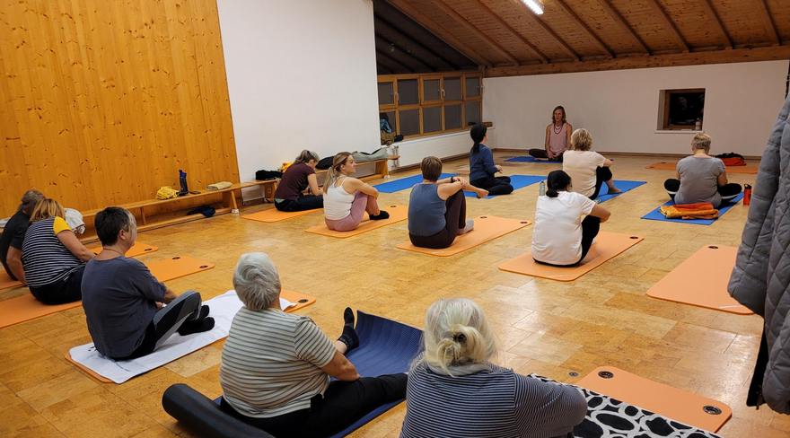 Das Bild zeigt mehrere Personen, die in einem beleuchteten Raum mit Schrägdach und Holzboden auf Yogamatten sitzen. 