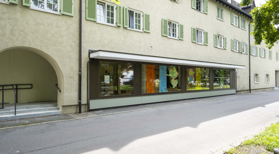 Das Bild zeigt die neue Jugendanlaufstelle "JUB" in Bregenz Rieden von außen. Es ist ein Haus im Stil der Südtirolersiedlungen zu sehen, unten sind mehrere Schaufenster mit bunten Aushängen bestückt. 