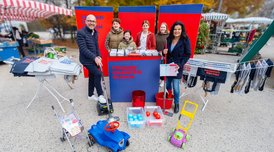 Das Bild zeigt den Stand des Frauenservice der Stadt Bregenz, der auf dem Kornmarktplatz aufgestellt ist. Daneben sind ein Wäscheständer und ein Bügelbrett zu sehen, davor verschiedenes Kinderspielzeug. Bürgermeister Michael Ritsch und Vizebürgermeisterin Sandra Schoch stehen neben städtischen Bediensteten vor dem Stand. 