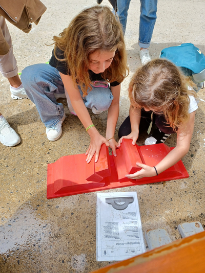 Man sieht zwei Mädchen am Boden am Basteln mit einem roten Werk. Vor ihnen liegt eine Anleitung am Boden, sie blicken runter. 