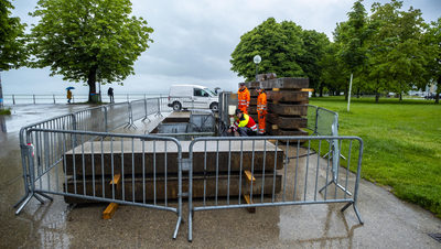 Auf dem Foto sieht man, wie eine Hochwasserpumpe im Boden verlegt wird. Es sind Bauhofermitarbeiter in orangen Anzügen zu sehen. Im Hintergrund der See. 