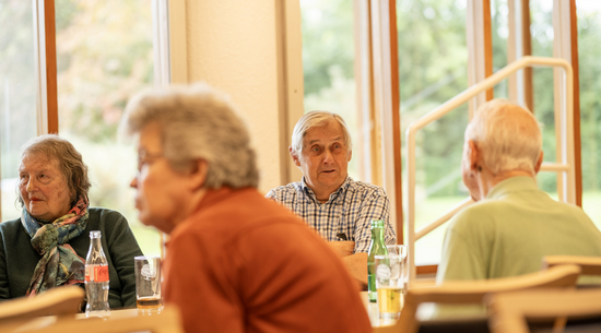 Das Bild zeigt eine Veranstaltung des Seniorenclubs im Tschermakgarten. Zu sehen sind vier Senior:innen, die zusammen an einem Tisch sitzen und teilweise der:dem Vortragenden lauschen, während zwei Personen sich unterhalten.