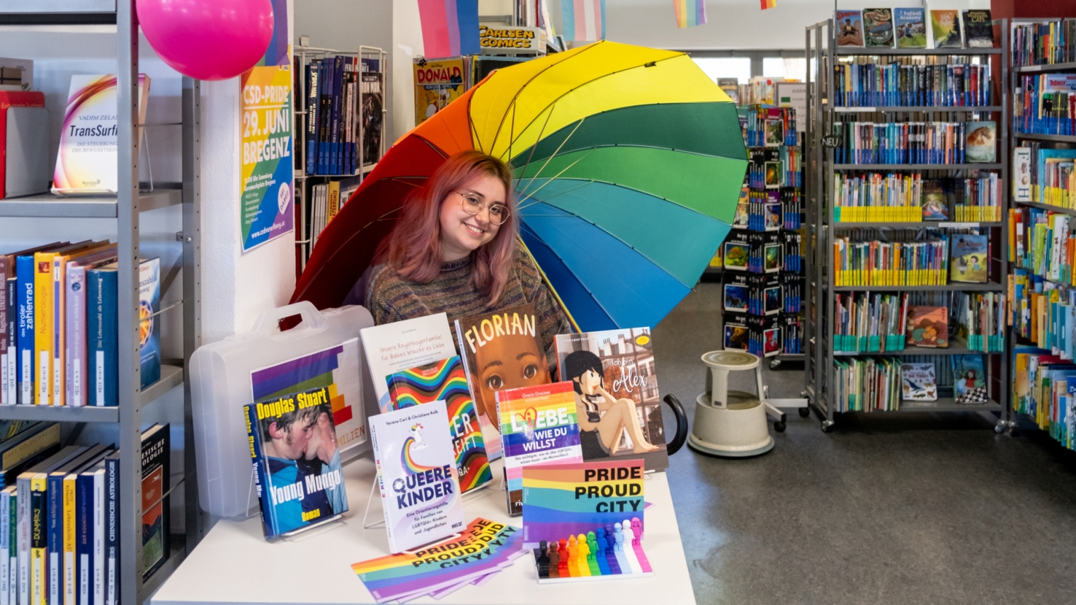 Das Bild Zeigt Maria Götz vom Team der Bregenzer Stadtbücherei an einem Bücherinfotisch mit ausleihbaren queeren Büchern.