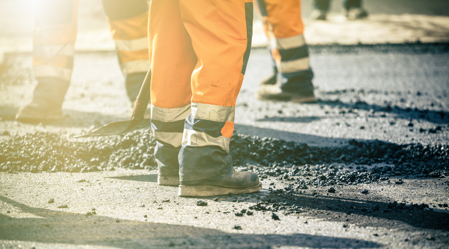 Das Bild zeigt eine Straße, die gerade geteert wird. Darauf zu sehen sind die Beine mehrerer Straßenarbeiter, zu erkennen an ihrer orangen Uniform mit Reflektorstreifen und Arbeitsschuhen.