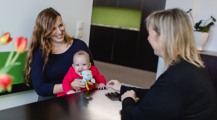 Diese Abbildung zeigt eine Mutter, welche mit ihrem Baby bei der Elternberatung ist. Sie sitzen alle gemeinsam an einem Tisch.