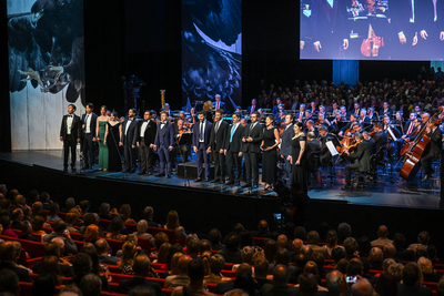 Auf diesem Bild sieht man eine Gruppe von Sängern in formeller Kleidung auf einer Bühne stehen. Hinter ihnen spielt ein großes Orchester. Im Publikum sitzen viele Zuschauer.