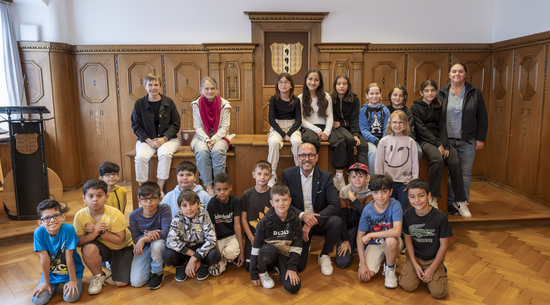 Das Bild zeigt die Schulklasse 3a der Volksschule Schendlingen beim Besuch im Bregenzer Rathaus. Die Kinder sitzen im Stadtvertretungszimmer um einen hölzernen Tisch zusammen mit ihren Lehrer:innen und Bürgermeister Michael Ritsch.