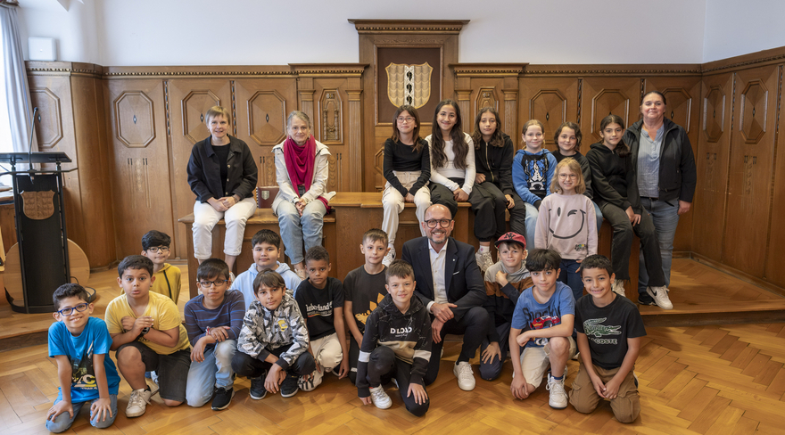 Das Bild zeigt die Schulklasse 3a der Volksschule Schendlingen beim Besuch im Bregenzer Rathaus. Die Kinder sitzen im Stadtvertretungszimmer um einen hölzernen Tisch zusammen mit ihren Lehrer:innen und Bürgermeister Michael Ritsch.