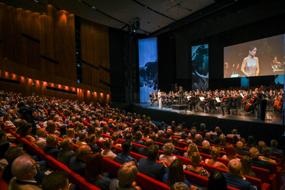 Auf diesem Bild sieht man einen großen, gut besuchten Konzertsaal. Das Publikum sitzt in roten Sitzen und schaut auf die Bühne, wo ein Orchester spielt. Im Hintergrund sind Projektionen und Bildschirme zu sehen.