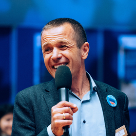 Man sieht ein Portrait von Dr. Heinz Fuchsig mit einem Mikrofon in der Hand am lächeln.