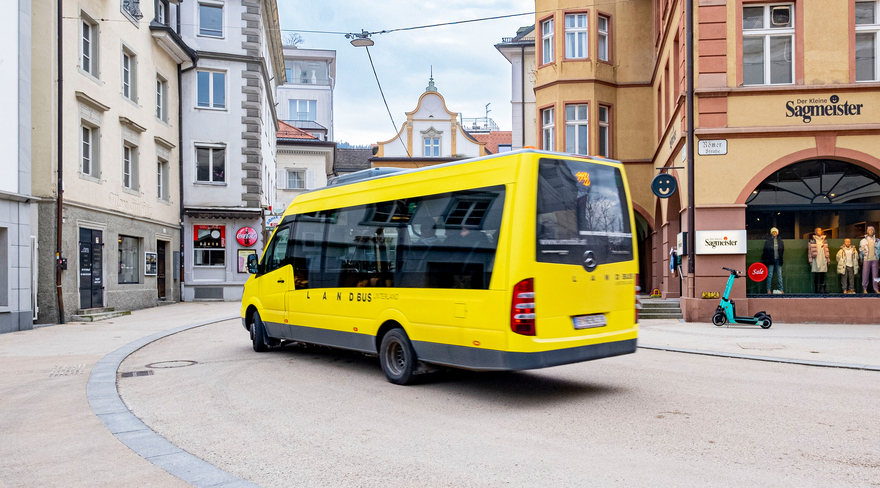Auf dem Bild ist ein kleiner, gelber Landbus von hinten zu sehen, der gerade von der Römerstraße abbiegend in die Kirchstraße unterwegs ist. 