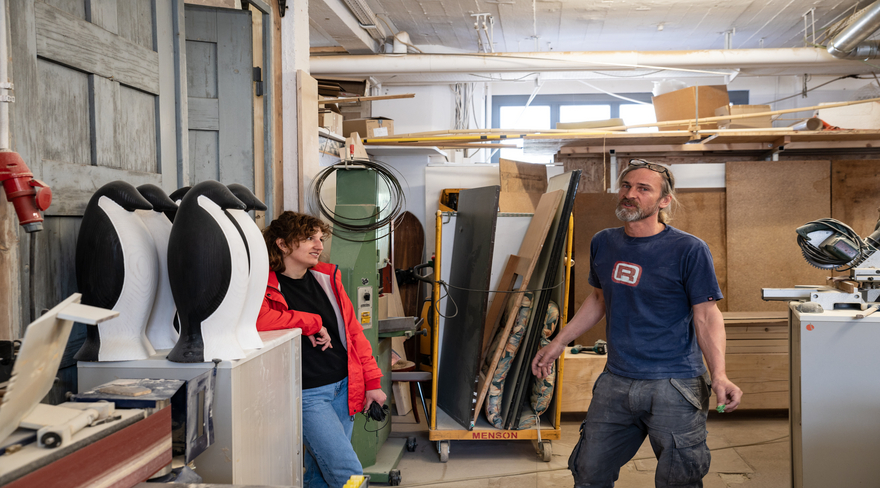 Eine Frau und ein Mann sind in einer Schreinerwerkstatt. Im Hintergrund sind Werkzeuge, Holzstücke und Arbeitsmaterialien zu sehen. Daneben stehen drei große, schwarz-weiß Holzskulpturen in Form von Pinguinen. Die Frau trägt eine rote Jacke und der Mann ein blaues T-Shirt.