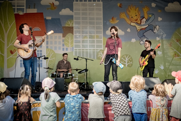 Das Bild zeigt die Künstler Michaela und Jimmy auf einer Bühne vor einem Kinderpublikum. Im Hintergrund sind zwei Musiker an der Gitarre und am Schlagzeug auf der Bühne.