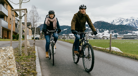 Das Bild zeigt einen Mann und eine Frau auf dem Fahrrad, die mit Fahrradhelm ausgestattet eine Straße entlangfahren. Am Wegesrand ist teilweise Schnee zu sehen. 