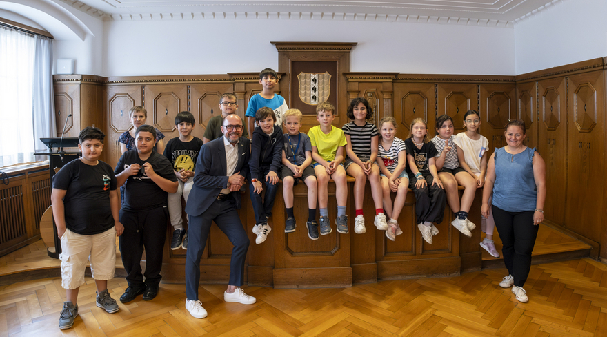 Das Bild zeigt die Schulklasse 3c der Volksschule Schendlingen beim Besuch im Bregenzer Rathaus. Die Kinder sitzen im Stadtvertretungszimmer um einen hölzernen Tisch zusammen mit ihren Lehrer:innen und Bürgermeister Michael Ritsch.