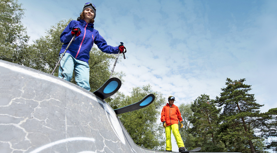 Diese Abbildung zeigt einen Jungen und ein Mädchen, welche in Winterklamotten mit Ski und einem Snowboard auf Rampen stehen.