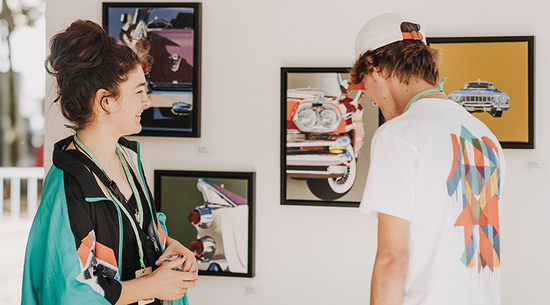 Das Bild zeigt zwei junge Personen, die Bilder betrachten, die auf einer weißen Wand im Hintergrund aufgehängt sind. Beide lachen dabei. 