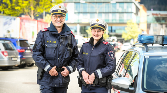 Auf dem Foto ist ein Polizist und eine Polizistin zu sehen wie sie in die Kamera lächeln. Hinter ihnen ist ein Parkplatz bei Tag. 