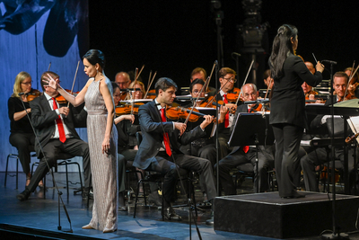 Auf diesem Bild sieht man eine Frau in einem glitzernden Kleid, die auf einer Bühne vor einem Orchester singt. Das Orchester spielt Geigen und andere Instrumente. Im Hintergrund sind große Projektionen zu sehen.