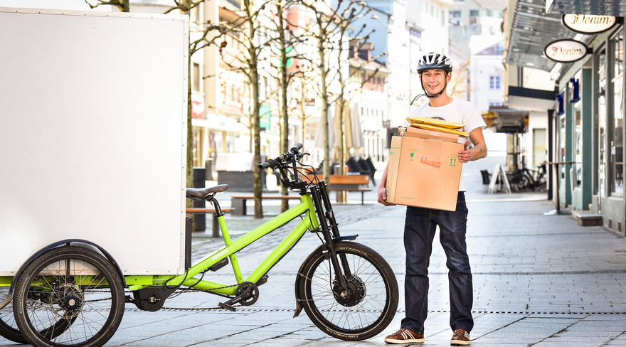 In dieser Abbildung ist ein Herr zu sehen, welcher mit dem Fahrrad und einer Box, die er den Bürger:innen liefert, in der Bregenzer Innenstadt fotografiert wurde. 