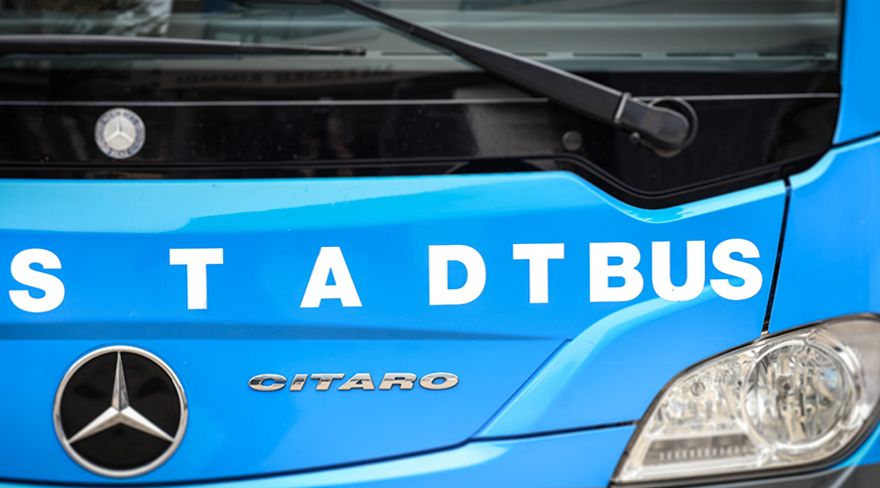 Auf diesem Bild sieht man einen Stadtbusfahrer in einem blauen Stadtbus in Bregenz. Der Bus fährt auf einer Straße in einem Wohngebiet. Die Umgebung ist grün und ruhig.