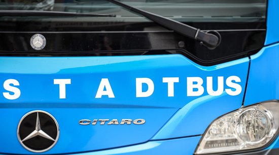 Auf diesem Bild sieht man einen Stadtbusfahrer in einem blauen Stadtbus in Bregenz. Der Bus fährt auf einer Straße in einem Wohngebiet. Die Umgebung ist grün und ruhig.