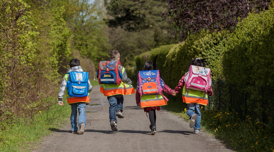 Diese Abbildung zeigt vier Schulkinder mit Schultaschen, welche Warnwesten tragen und zusammen ihren Schulweg gehen. 
